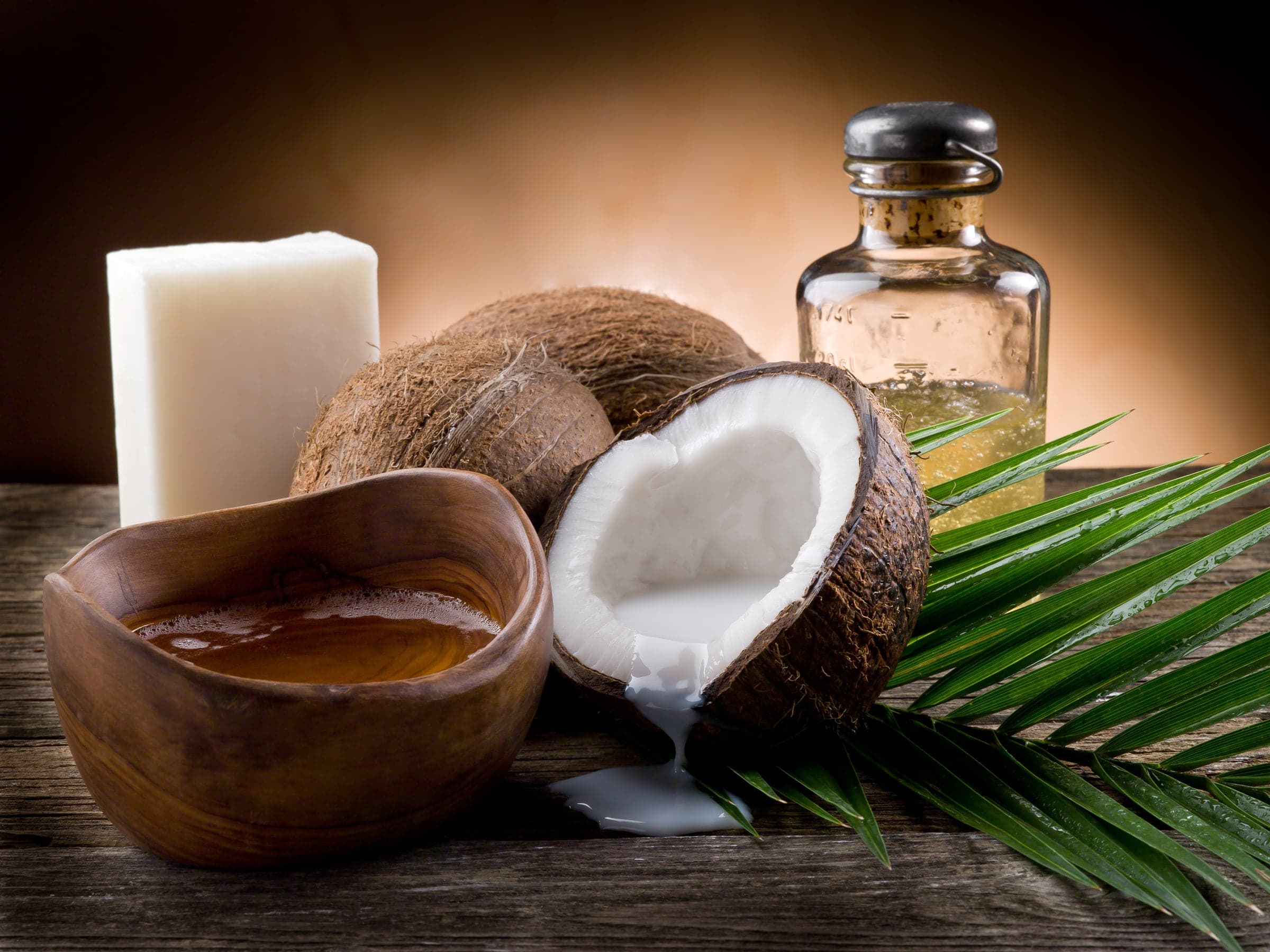 Coconut halves and a bowl of oil on dark wood with a palm leaf