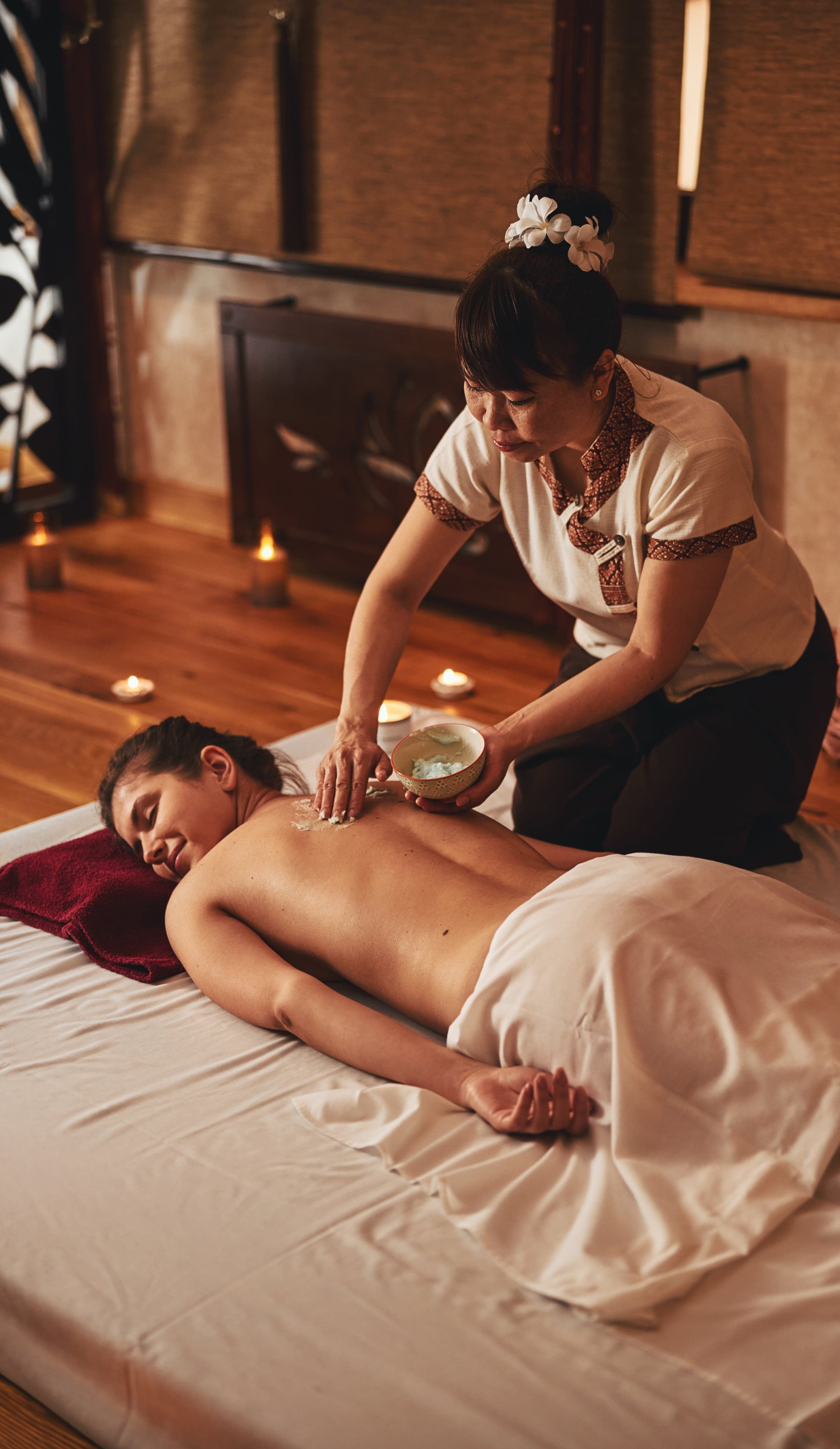 Hot coconut oil massage — a therapist applies a warm blend from a coconut-shell bowl to the back of a guest lying on a mat, candlelight around