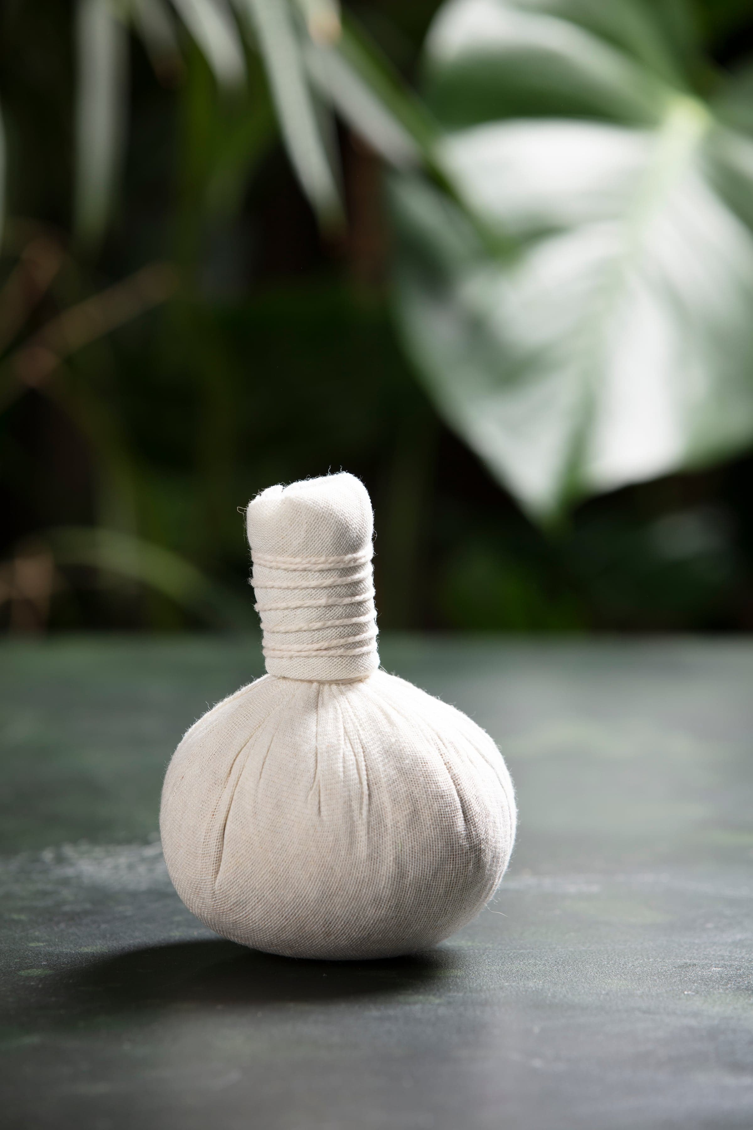 A Thai herbal compress in linen on dark stone, green foliage behind